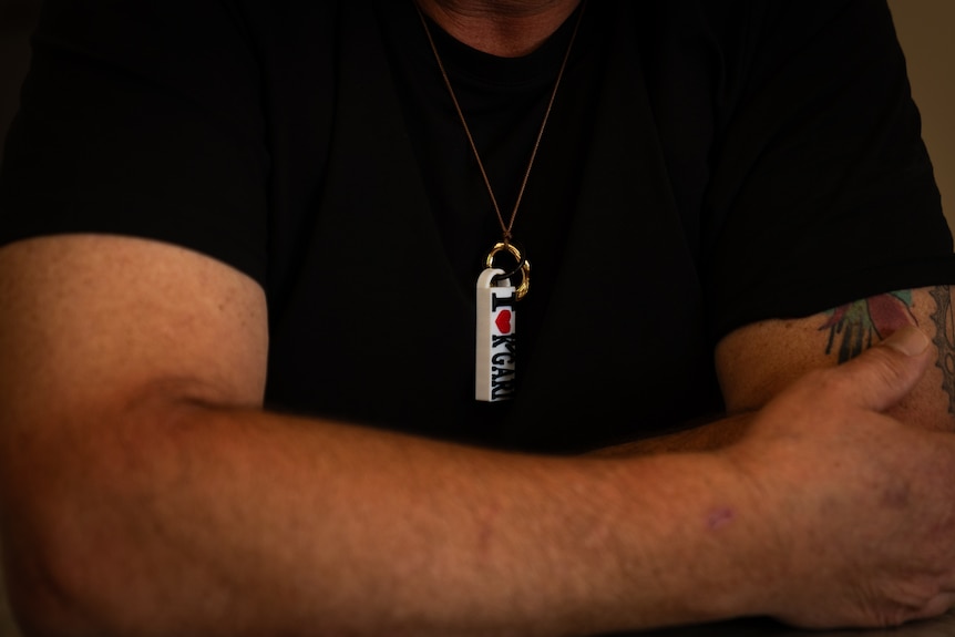 Man in black tee shirft with arms folded wearing a necklast with a pendant reading I love K'Gari