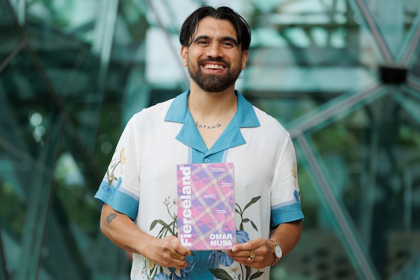 Omar Musa, a dark-haired man with a short beard, wearing a short-sleeved button-down shirt and holding a book in his hands.