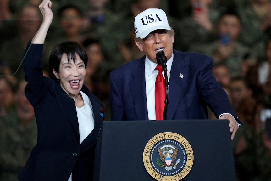 Donald Trump speaks into a podium next to Sanae Takaichi, who cheers with her arm in the air.