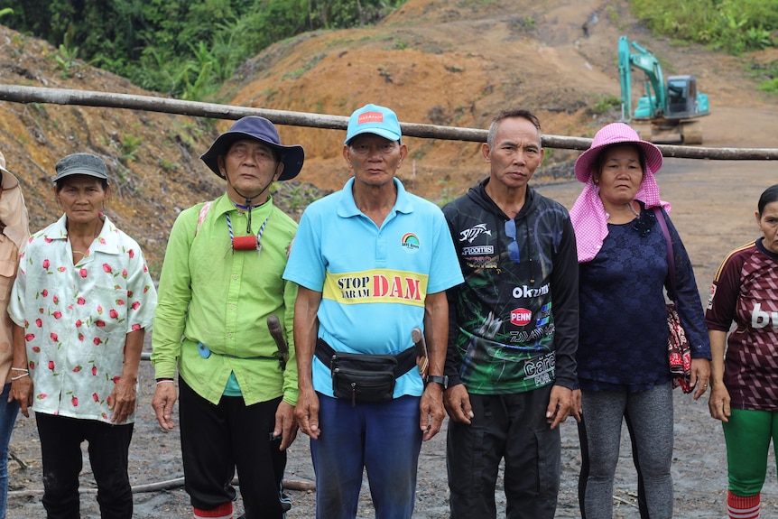 A line of people with sad faces stand in front of logging machinery