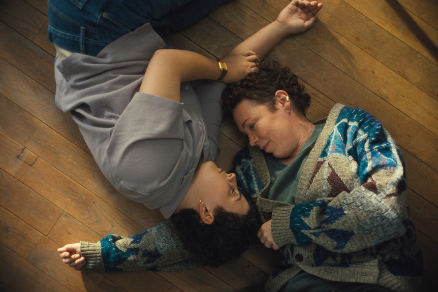 A young person with dark, short, curly hair lies on the floor, arms toward mother, who lies next to them wearing knit jumper