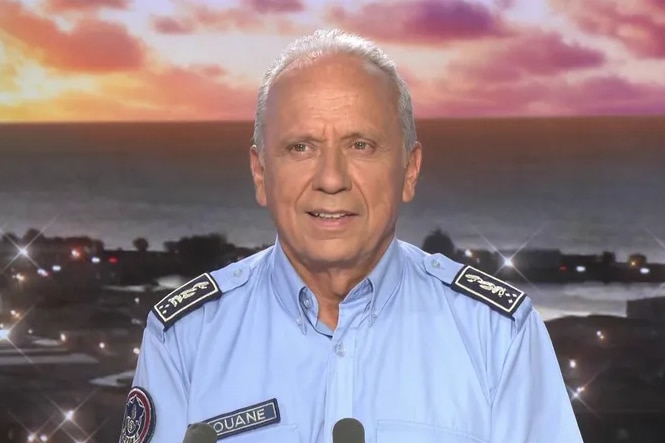 A grey-haired man in a uniform sits in what appears to be a TV studio with a backdrop of a port.
