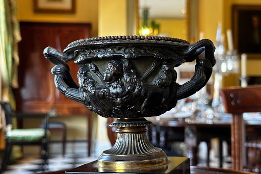 A small dark urn with decorative handles and faces in a formal room.
