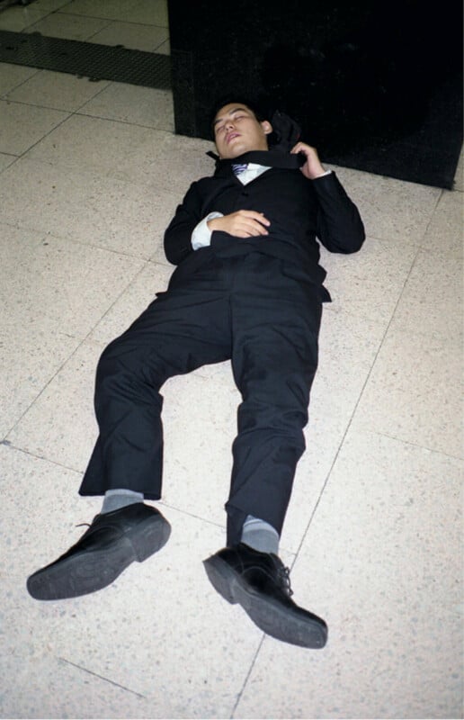 A man in a black suit and tie is lying on his back on a tiled floor, eyes closed, with one hand resting on his chest and the other behind his head.