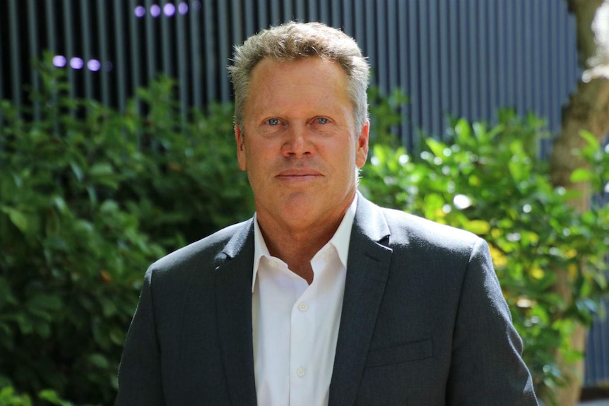 A close up of Gavin Hegney standing outside in the sunshine with green shrubs and a grey wall behind him.