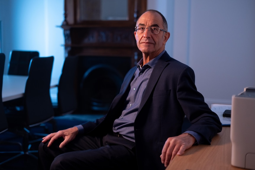 A man wearing a dark suit and shirt is sitting and looking directly at the camera. Behind him is a board room table and chairs