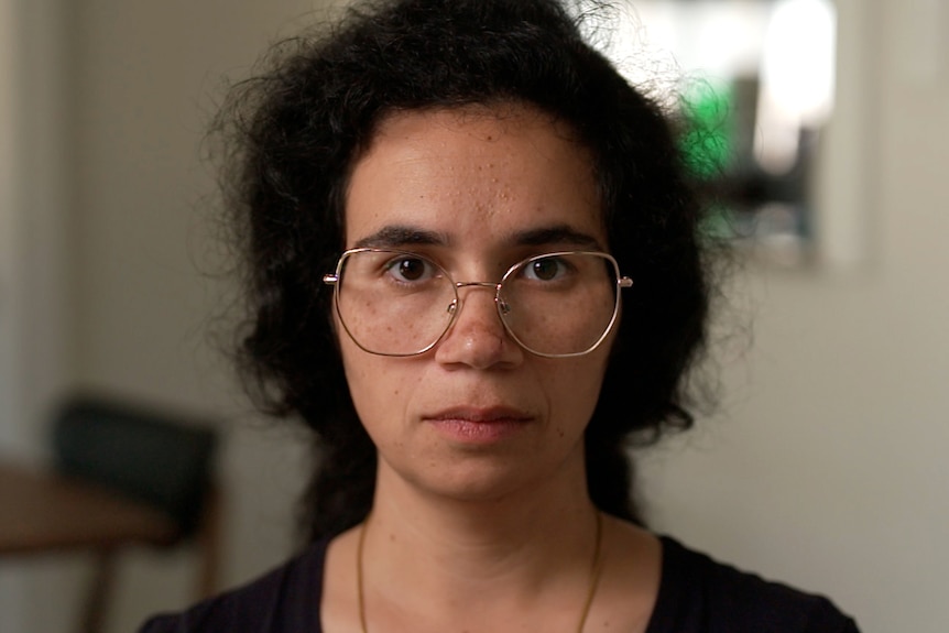 A close-up portrait of a woman who is looking intently at the camera with a serious expression. She wears glasses.