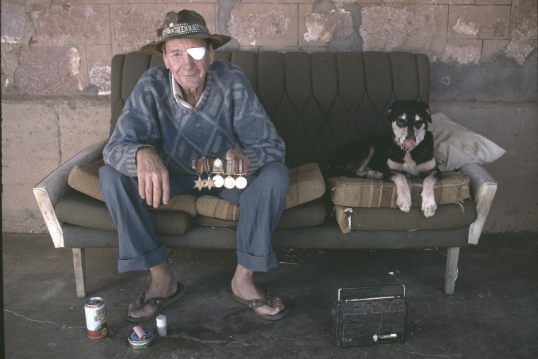 An old prospector with an eye patch sits on a couch with a dog. 