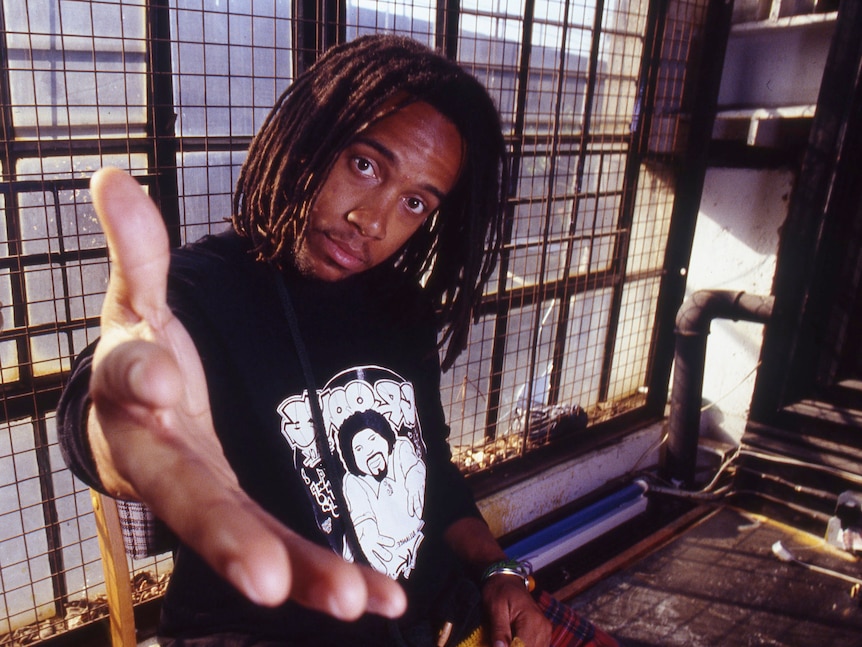 Man with dreadlocks in a warehouse wearing a black shirt and holding his hand out to the camera.