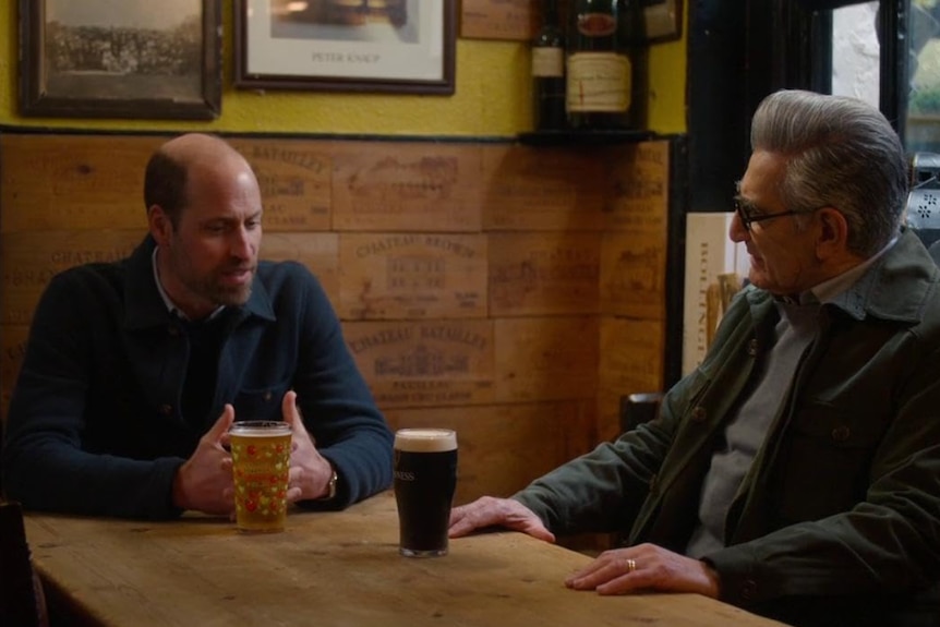 Prince William sits at a table wtih a beer while sitting across from Eugene Levy.
