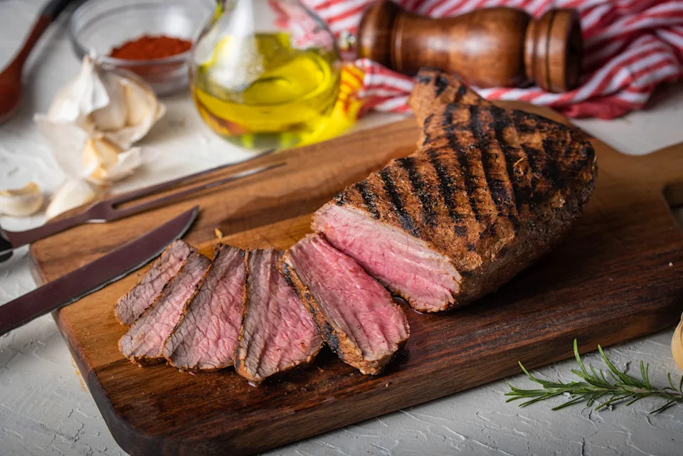 Grilled steak sliced on a wooden cutting board, surrounded by olive oil, garlic, and spices