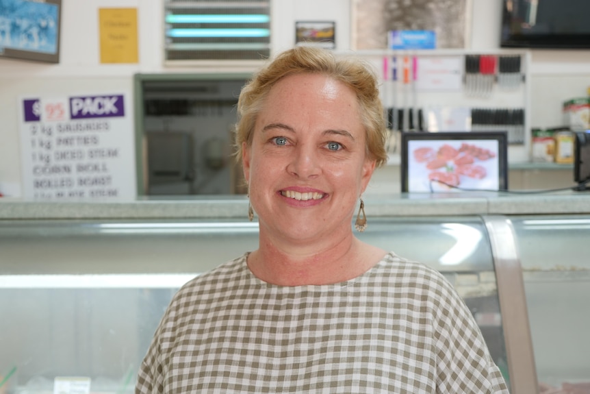 Woman in checked shirt standing in front of meat counter at butcher shop smiling at camera.