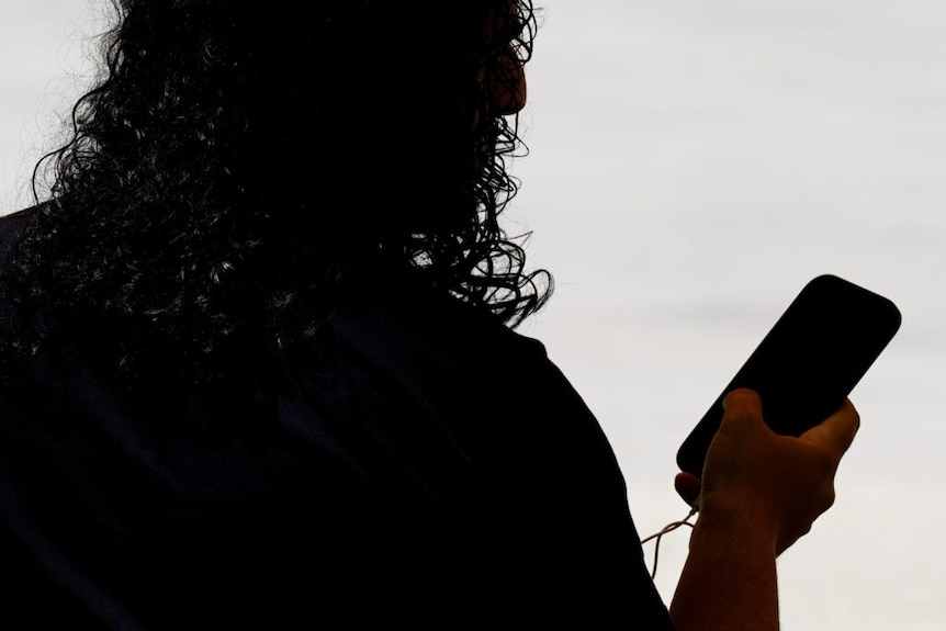 A dark silhouette of a man with curly hair looking to the right of frame while holding a mobile phone with a single hand