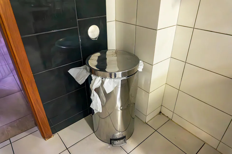 Metal trash can with a pedal is placed in a tiled corner, with paper peeking out from under the lid