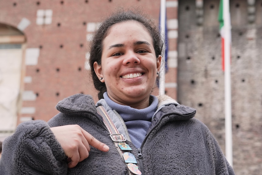 A woman smiles while pointing at an Olympic pin on her bag