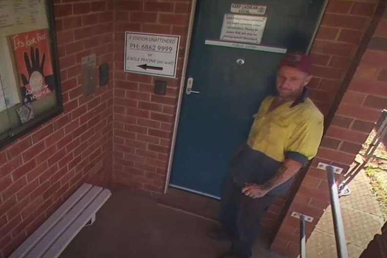 Man in yellow hi-vis shirt stands outside a closed door at a police station.