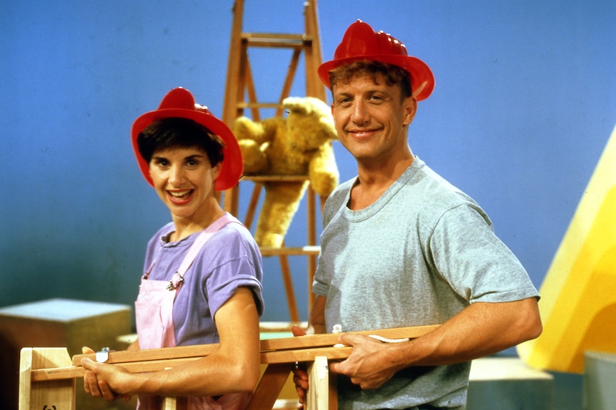 A TV still of Monica Trapaga, mid-20s, and Simon Burke, 30, smiling brightly in fire helmets, carrying an easel together.