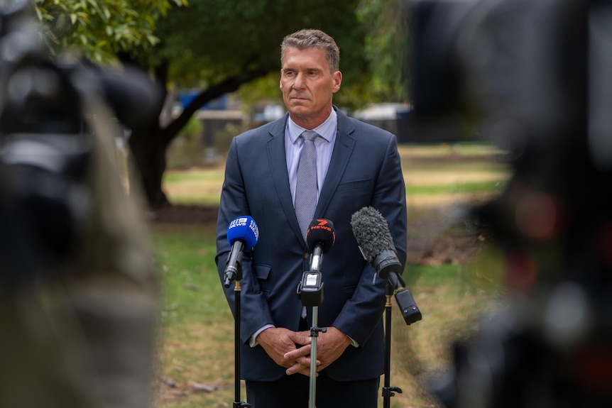 A man in a suit stands in front of three microphones with a neutral expression