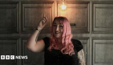 Debbie Todd has pink wavy hair. She is wearing red lipstick and a black top as she points to a light bulb shining above her head. Her right arm is covered in tattoos.