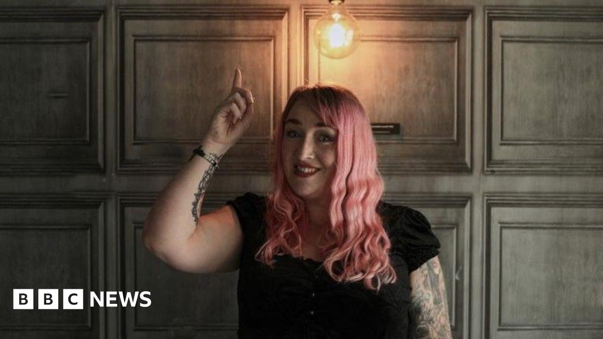 Debbie Todd has pink wavy hair. She is wearing red lipstick and a black top as she points to a light bulb shining above her head. Her right arm is covered in tattoos.