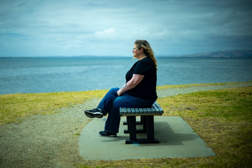 A woman sits on a park bench with the sea in the background.