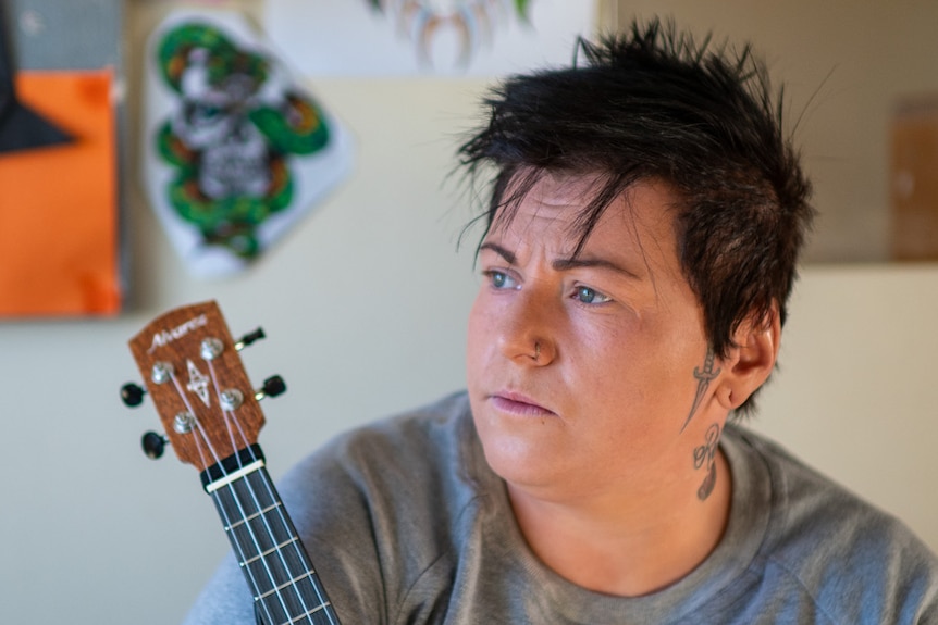 Fabiann looks to the left with a concerned expression while holding a ukulele and sat on her bed in her cell room.