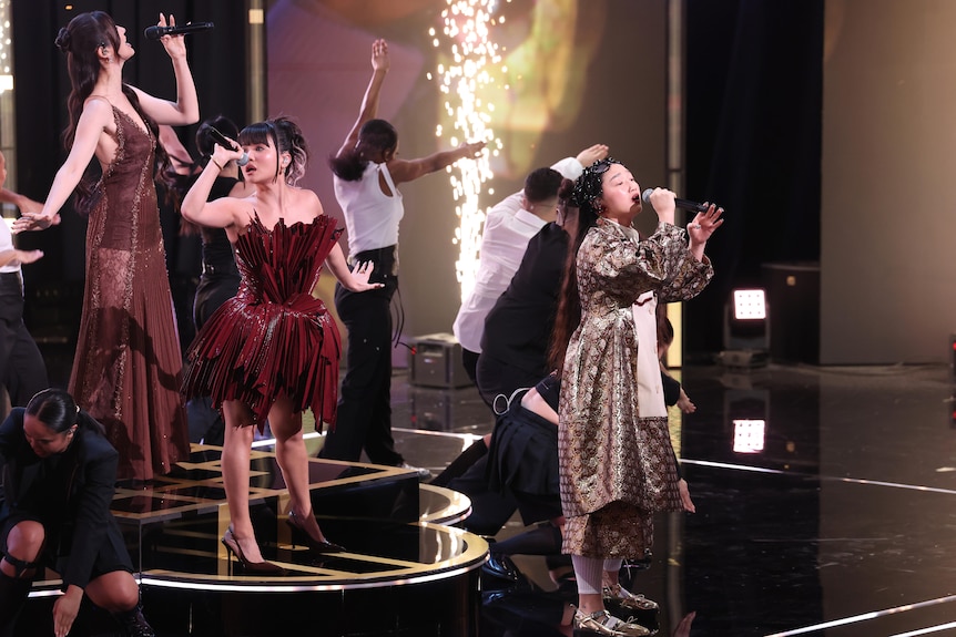 Three women sing on stage with backup actors and pyrotechnics