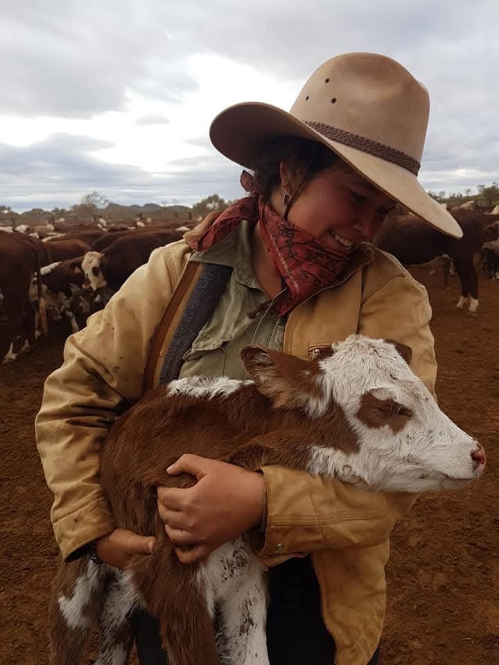 A smiling woman in a broadbrimmed hat and bandana holds up a young calf.