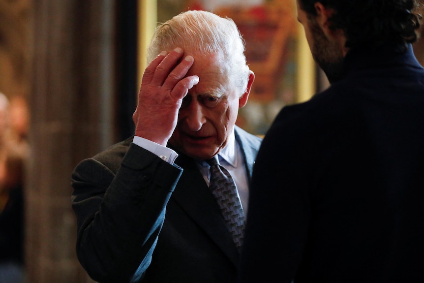 Britain’s King Charles reacts by holding his hand up to his head