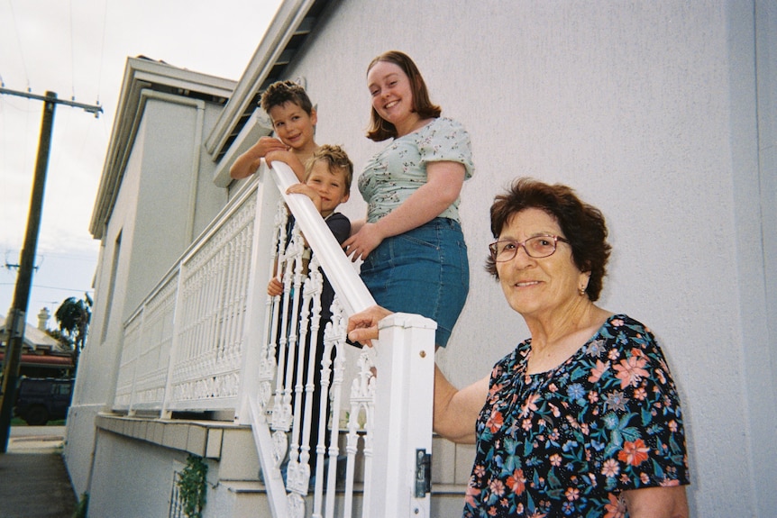 An older woman stands at the bottom of outdoor stairs, with a younger woman and two children standing above her.