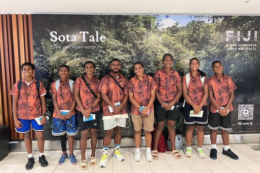 A group of young people in brown button shirts and different coloured shorts and shoes stand in a line to pose in an airport