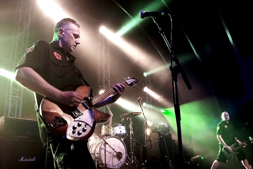 Two people play guitars on a stage with drums in the background