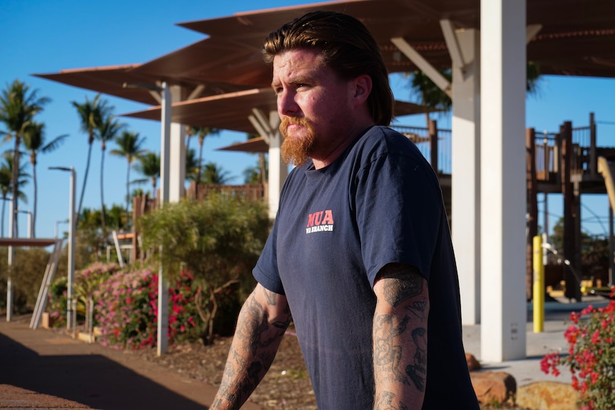 A man with long red hair and beard looks out at the horizon, leaning against a brick sea wall.