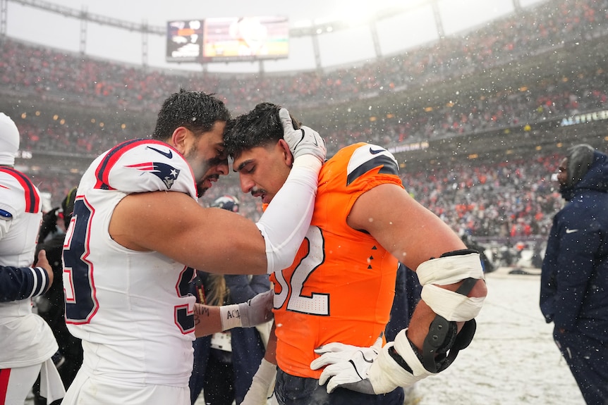 A man in a white Patriots football jersey touches foreheads with a man wearing an orange Broncos jersey with arms around him