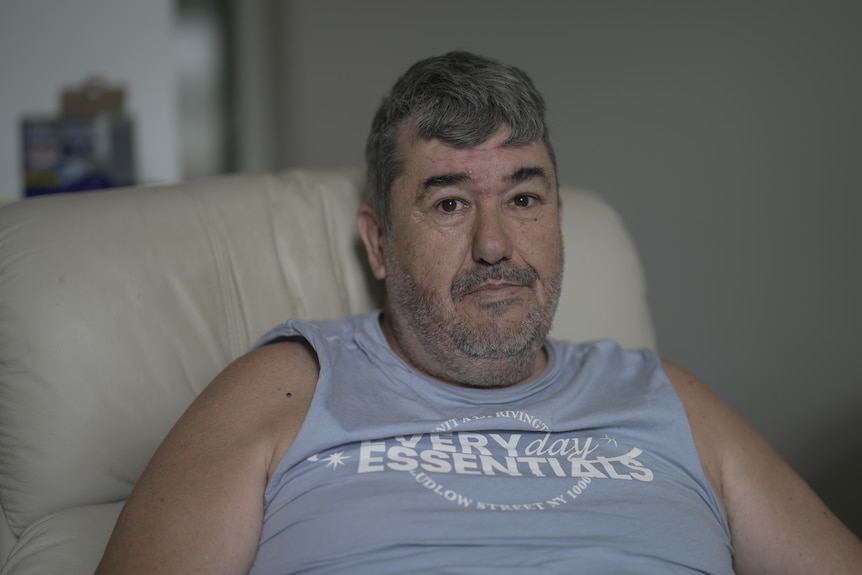 Man in a light blue tank top sitting on a white armchair.