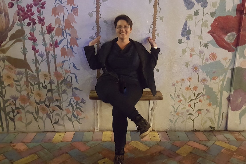 A smiling young woman with short dark hair and glasses, dressed in black, sits on a bench chair against a wall with artwork.