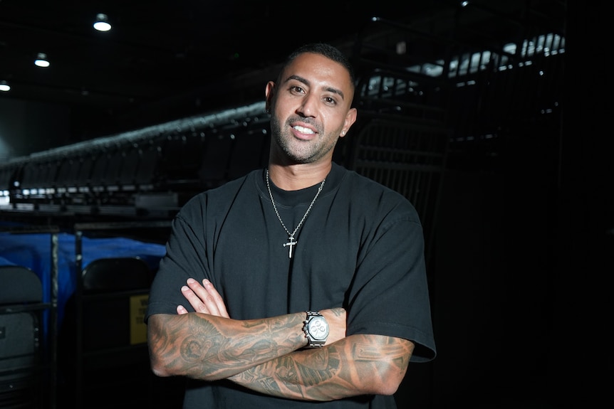 a man smiling at the camera, he has his arms crossed and is standing in an indoor sporting event space