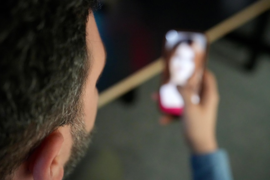 An anonymous man with dark hair and a dark beard seen from behind, looking at a blurred mobile phone with a woman's image.