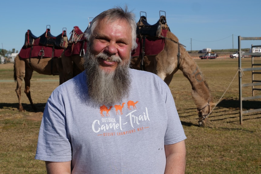 man with beard wearing camel shirt smiling, camel behind him