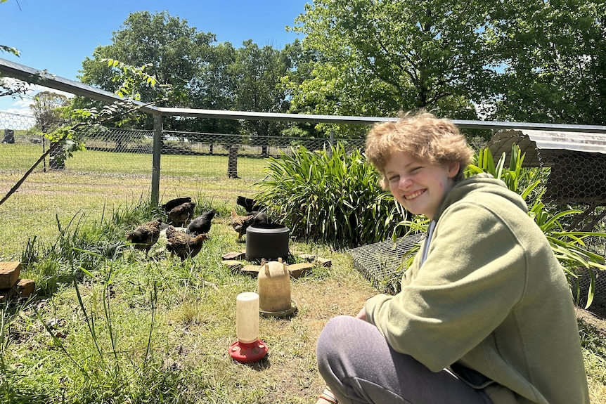 A blonde teenager in a green hoodie smiles, with several chickens in the background of a wired enclosure.