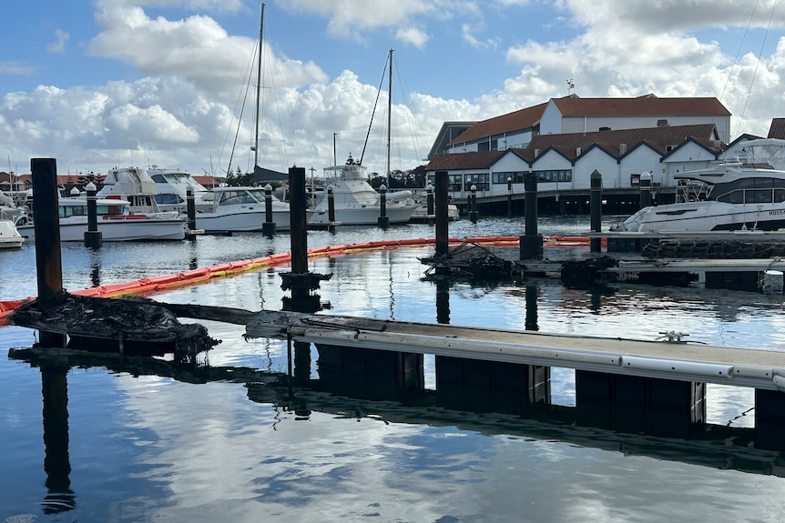 Empty berths at a boat harbour, with blackened remains of boats destroyed by fire.