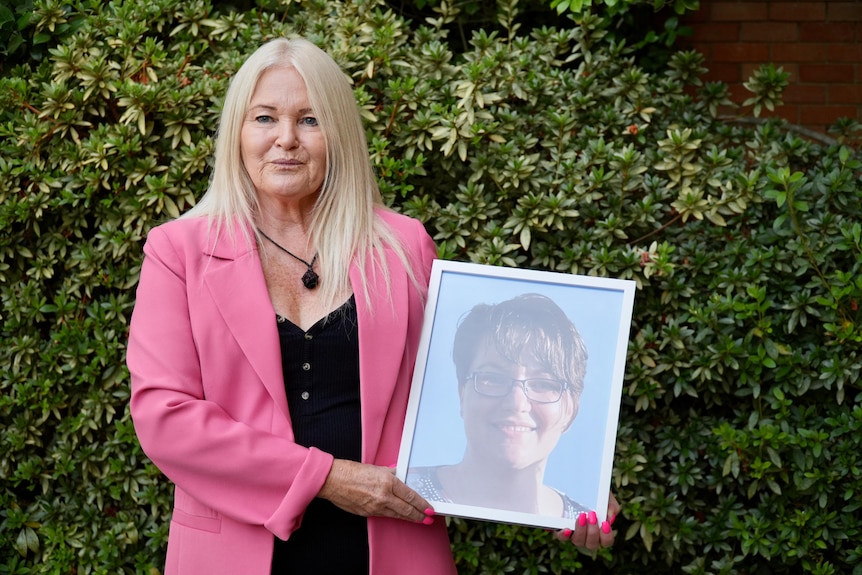 A woman with blonde hair, a pink jacket and a black shirt, holding a picture frame.