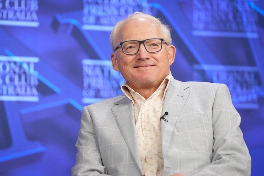 An older man in grey suit and black-rimmed glasses sits onstage at the National Press Club.