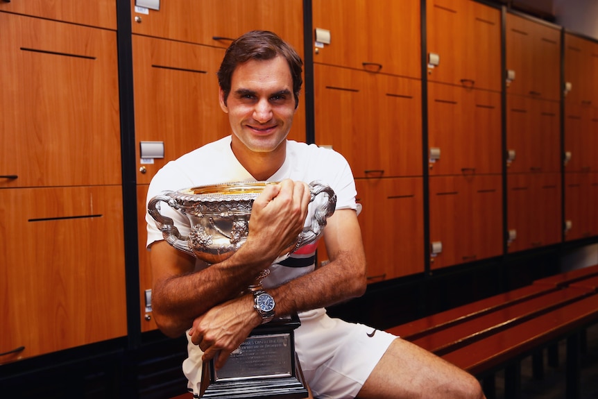A man in a white t shirt and shorts sits in a locked room wrapping both arms around a trophy.