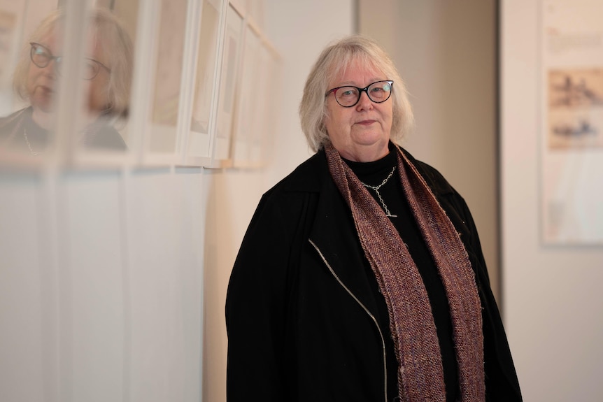 A woman in glasses stands in front of a wall of hanging artworks.