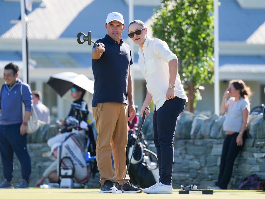 Surfer Kelly Slater and retired tennis star Ash Barty stand on a putting green as Slater points out a line for a putt.