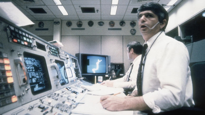 Two men in shirts and ties sit at a mission control desk. One looks past the camera, the other at a TV showing a forked cloud.