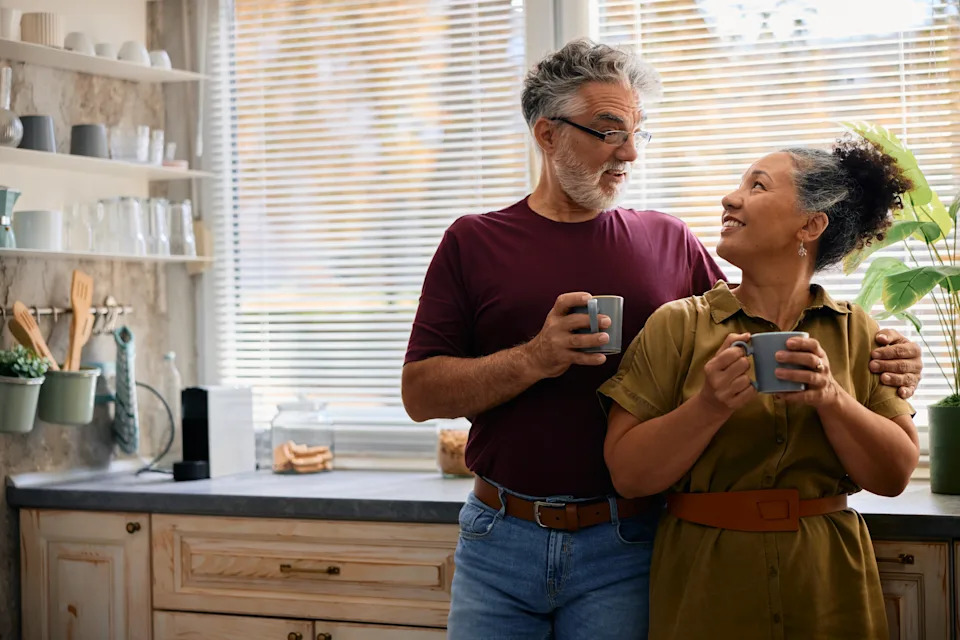 Happy senior couple enjoying their morning routine in a cozy kitchen, embracing and chatting while holding coffee cups, radiating love and togetherness in their relaxed atmosphere