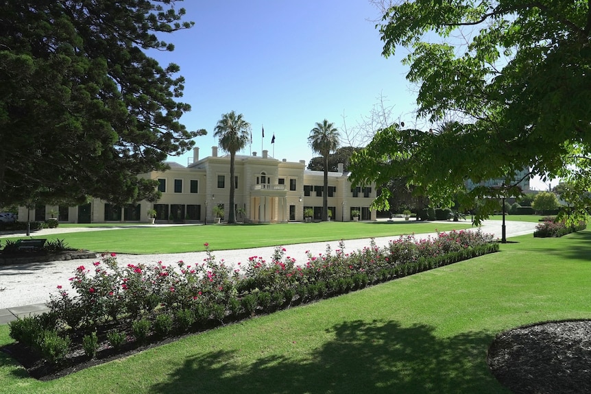 A large white building in the background. In the foreground are rows of pink flowers, green grass and trees.