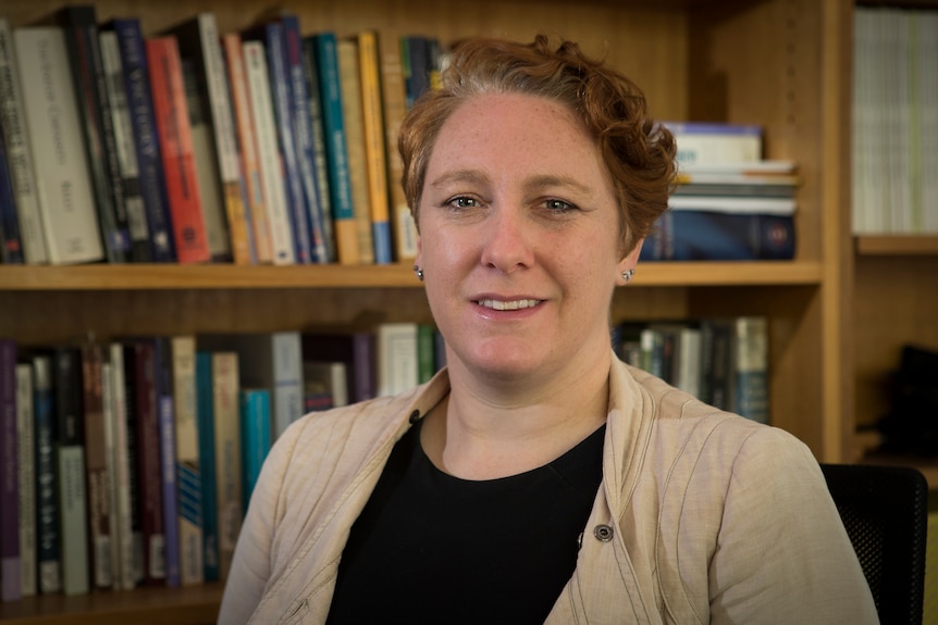 A woman with short, wavy hair smiles. A bookcase is behind her.